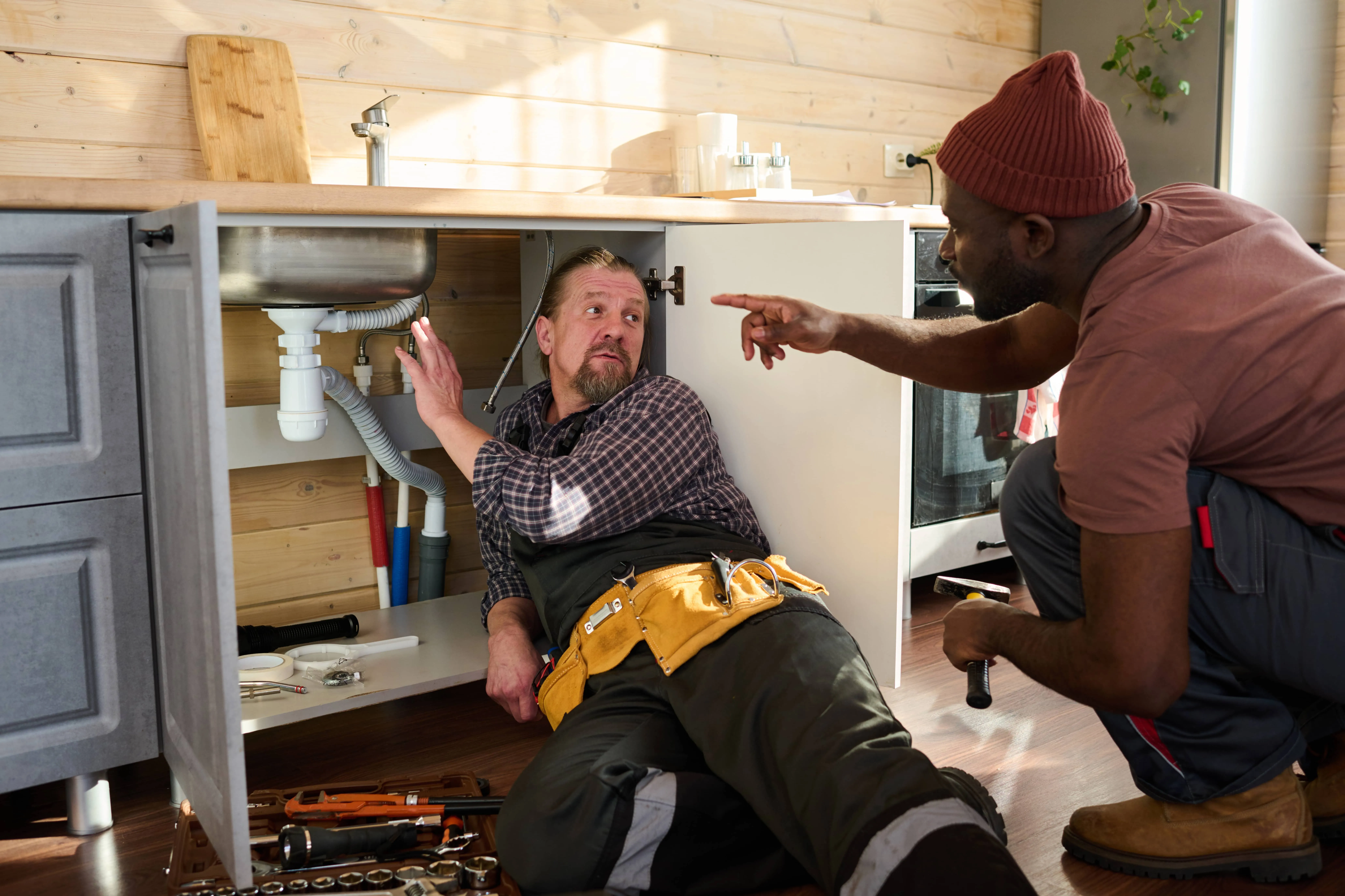 Two plumbers inspecting the drain pipe under a kitchen sink